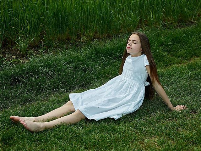 A young girl in a white dress relaxes on the grass in a peaceful outdoor setting, eyes closed enjoying the sunlight.