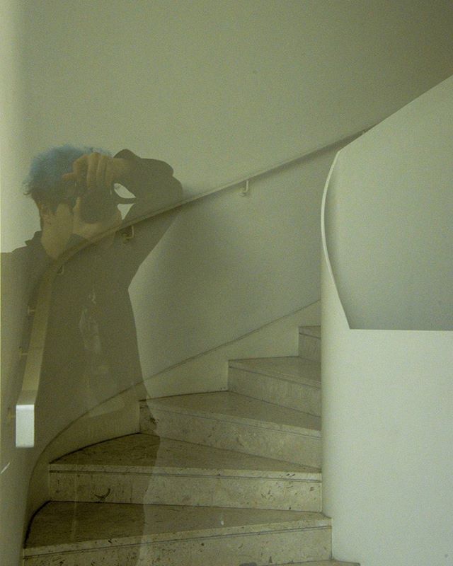 A person with blue hair takes a photo in a modern stairwell with a spiral staircase.
