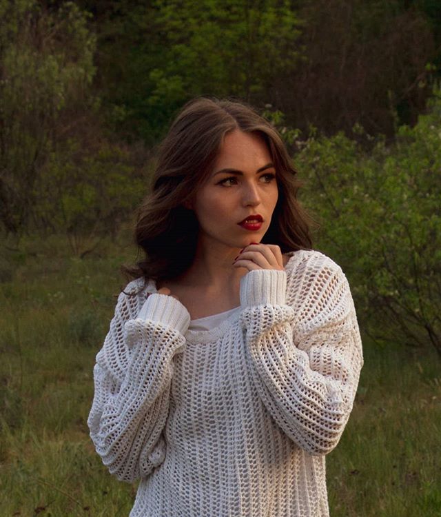 A brunette woman in a white sweater poses softly in a natural outdoor setting with a dreamy and contemplative mood.