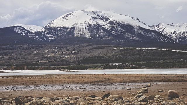 A landscape view of snow-capped mountains under a cloudy sky, with rocks and a barren field in the foreground.