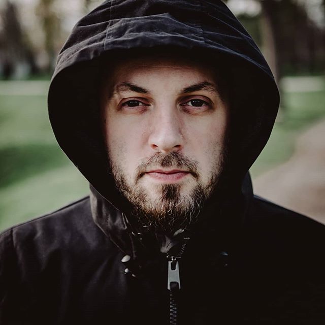 A man in a hoodie gazes directly at the camera in an outdoor setting, creating a moody and contemplative portrait.