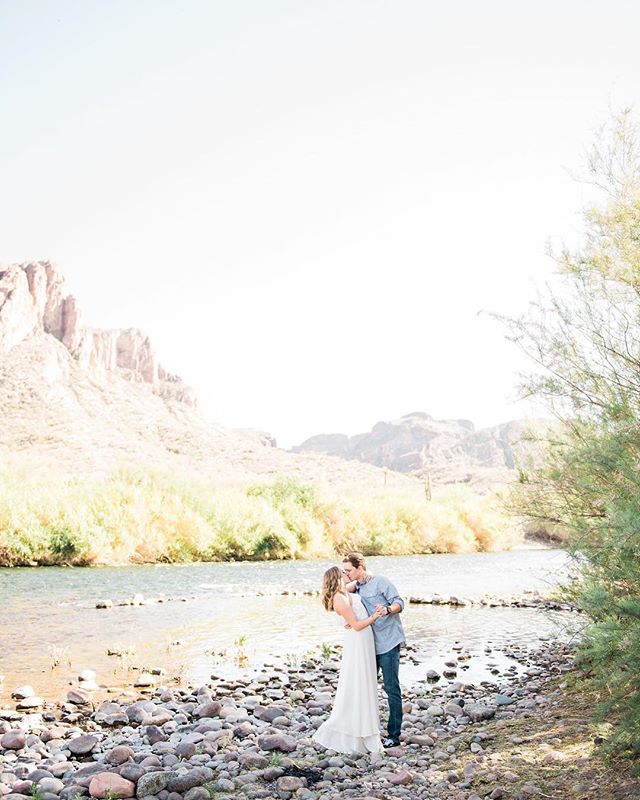 A couple in love share a kiss by the river with mountains in the background on a bright day.