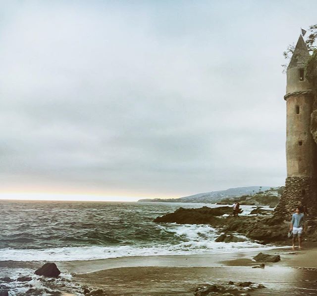 A man walks on a sandy beach next to a stone tower on a cloudy day by the ocean. 