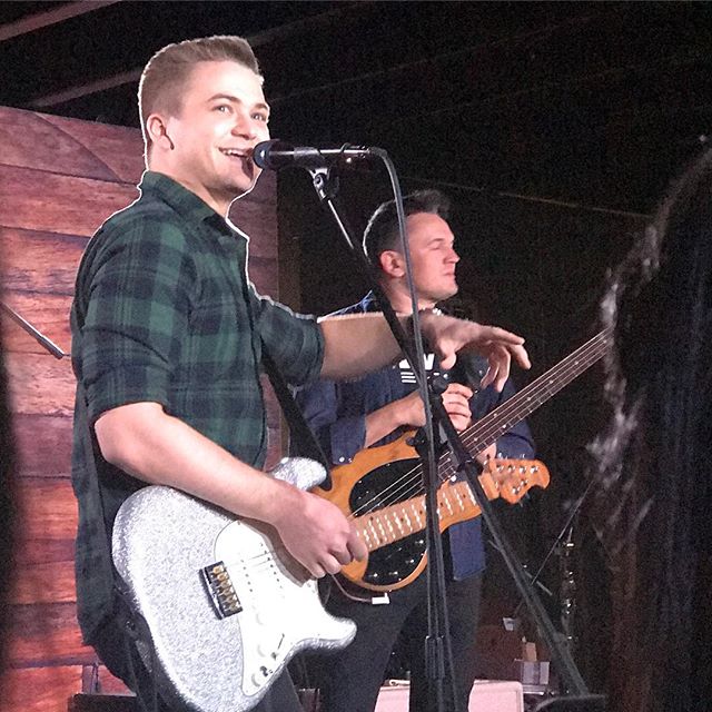Two musicians performing live on stage, one singing into a microphone while playing guitar.