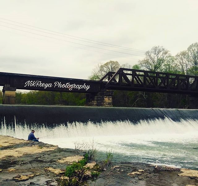 A man is fishing by a waterfall under a bridge, enjoying the calm and tranquil nature scene.