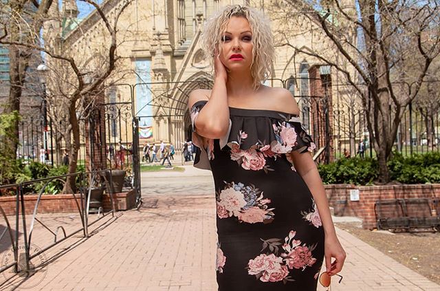 A stylish blonde woman poses in a floral dress in front of a church, exuding confidence and elegance.