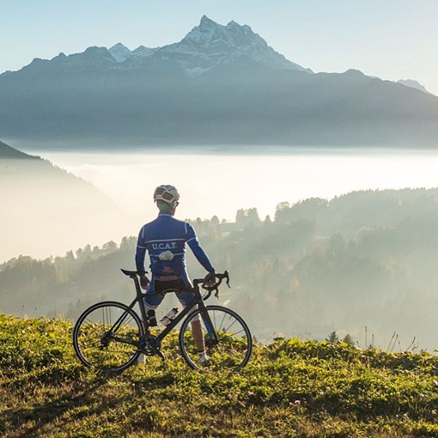A cyclist pauses on a hilltop with his bicycle, gazing at a scenic mountain range shrouded in mist.