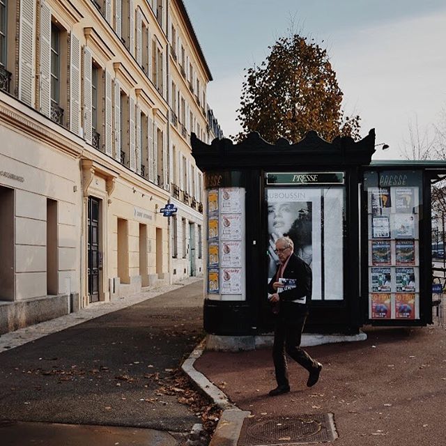 A man walks past a vintage newspaper stand on a quiet city street on a sunny day.