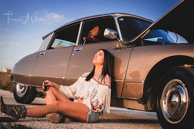 A woman relaxes against a vintage car on a sun-drenched road, holding sunglasses.