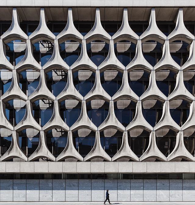 A person walks along a building with a unique, patterned concrete facade in an urban setting.