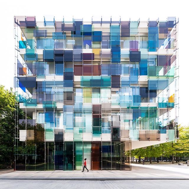 A modern building with colorful windows stands tall in the city, with a person walking on the sidewalk.
