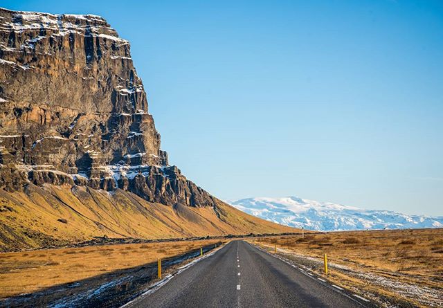 A scenic road stretches toward snow-capped mountains beneath a clear blue sky in Iceland.