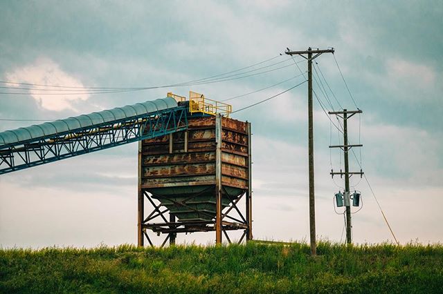 A silo with a conveyor belt connects to power lines against a cloudy sky in a rural setting.