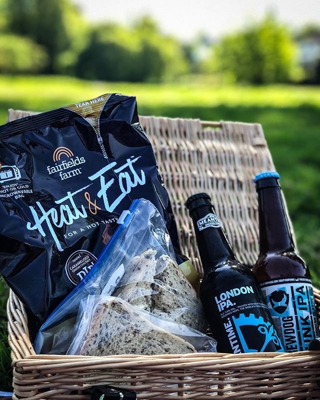 A picnic basket with beer, sandwiches, and chips set in a park for a relaxing outdoor dining experience.
