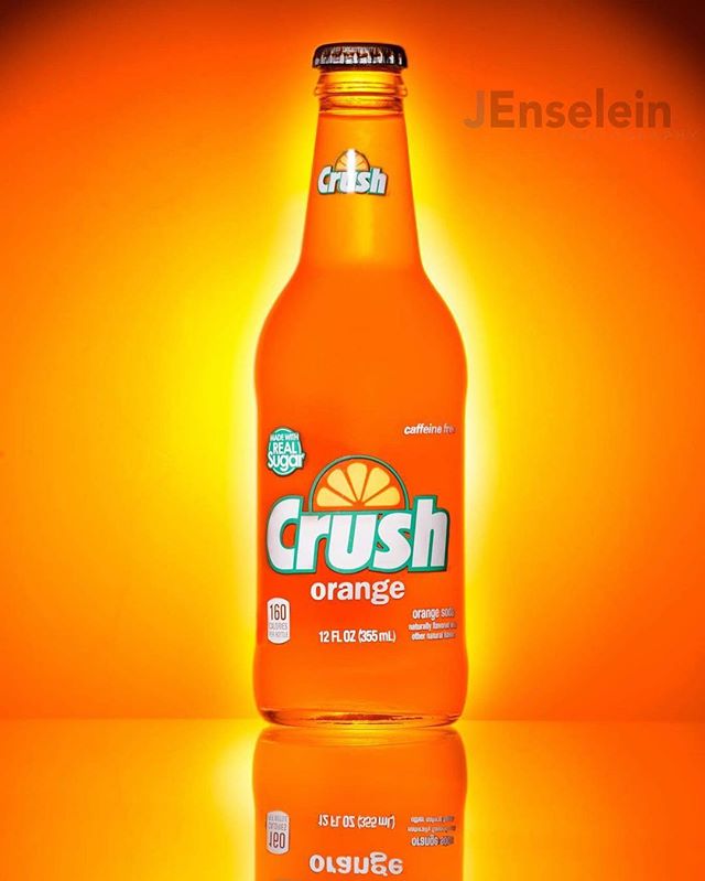 A bottle of orange Crush soda stands upright against an orange background, reflecting on a glossy surface.
