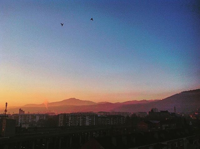 A serene cityscape at sunrise with mountains in the background and birds flying in the sky.