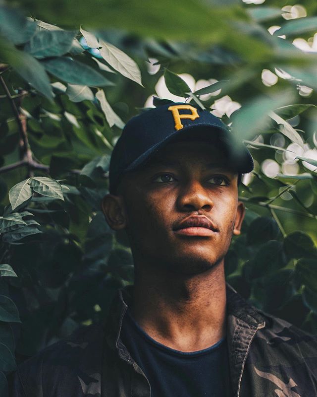 A man with a cap poses amongst green leaves, looking thoughtful.