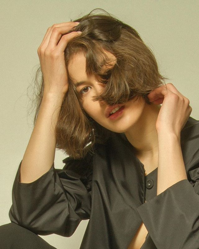 A brunette woman poses in a studio, creating a contemplative fashion portrait with soft lighting.