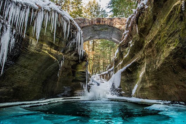 A scenic winter landscape of a canyon with a stone bridge, icicles, and a waterfall creating a tranquil and nature-filled vista.