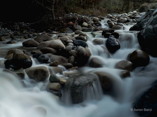 A river flows over smooth rocks in a serene natural setting, captured with a long exposure for a silky effect.