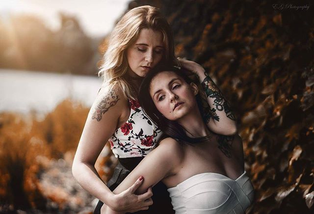 Two women with tattoos embrace tenderly outdoors in a romantic editorial style, with a soft, dreamy mood.