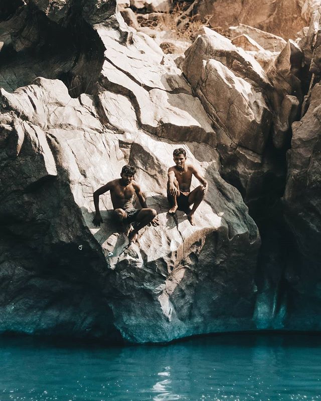 Two men sit on a large rock formation beside a river in a vintage-style editorial shot.