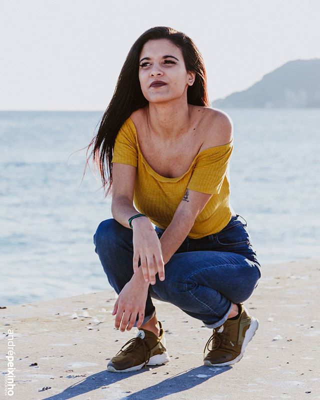 A woman squats on the beach in jeans and a yellow t-shirt, looking peaceful.