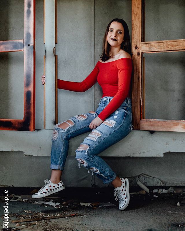 A young woman in a red top and ripped jeans sits on a ledge in an abandoned building.