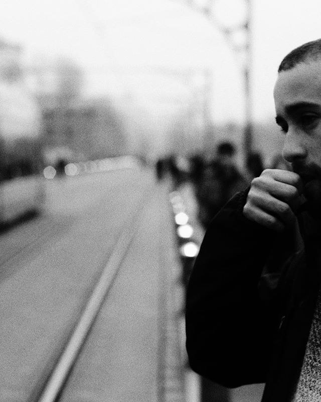 A thoughtful man with a beard stands on a bridge in a city, captured in black and white.