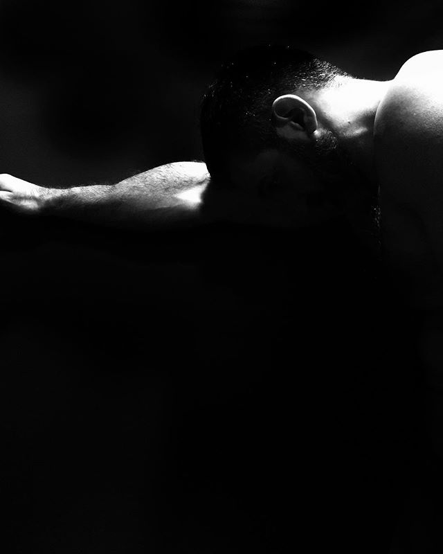 A muscular man rests his head on his arm in a dramatic, monochrome studio shot.