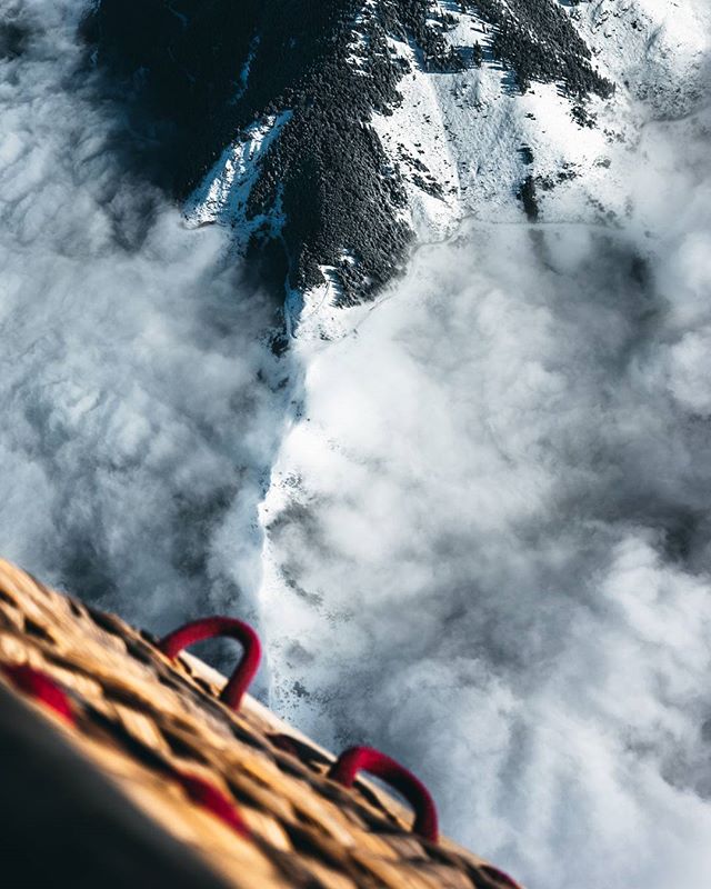 An aerial view shows snowy mountains through clouds from a hot air balloon basket.