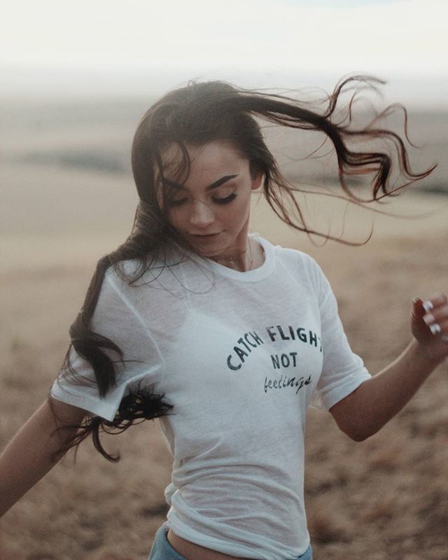 A young woman stands in a field with her hair blowing in the wind, wearing a t-shirt.