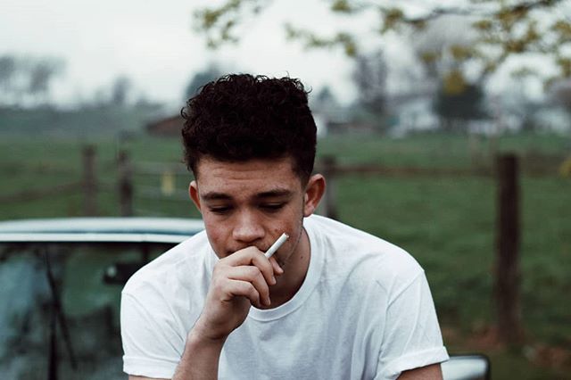 A young man in a white t-shirt sits smoking outside, lost in thought against a blurred rural backdrop.