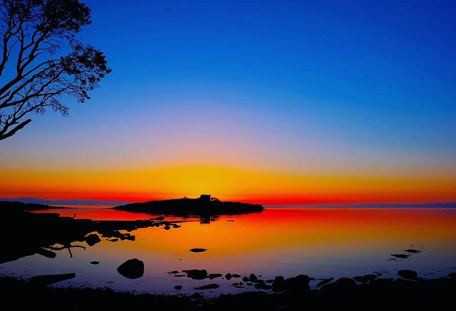 A tranquil sunset view of a calm lake reflecting vibrant orange and red hues, with a silhouetted island in the distance.