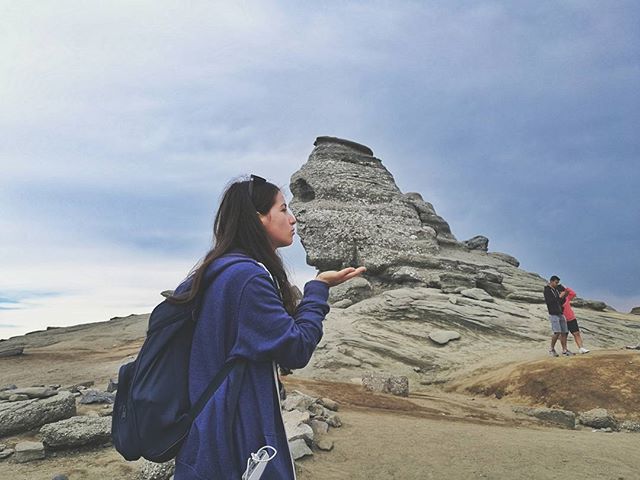 A woman blows a kiss towards a rock formation while a couple stands nearby on a mountain.