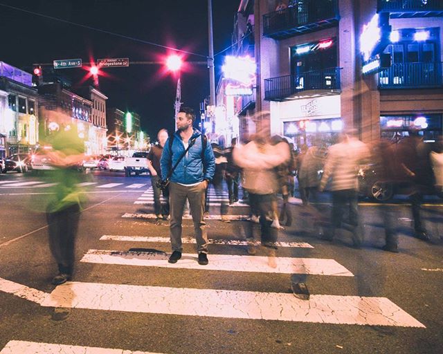 A man stands on a crosswalk amidst blurred people on a city street at night.