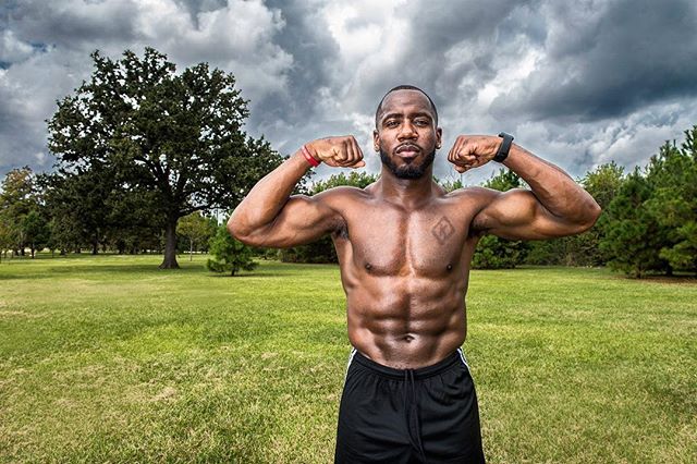A muscular man flexing in a park setting, showcasing fitness and strength under dramatic sky. 