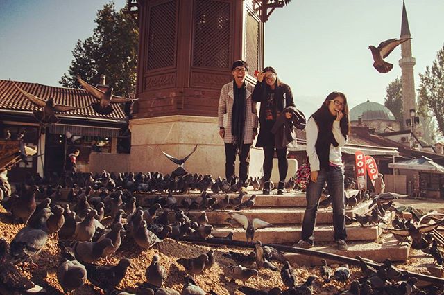 Three friends pose amid a flock of pigeons in a European city square with a minaret in the background.