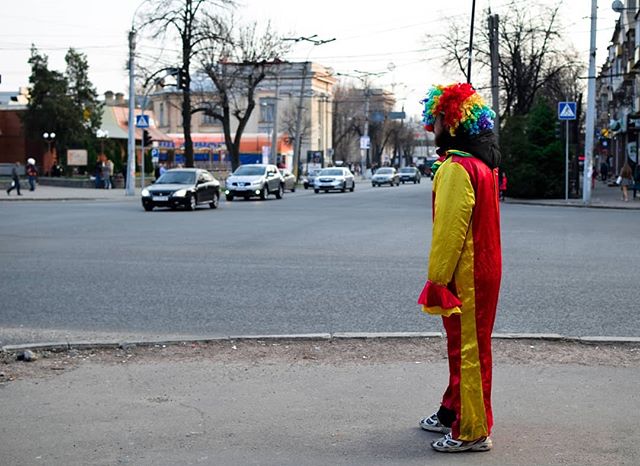 A clown in a colorful costume stands at a crosswalk on a city street with cars passing by.
