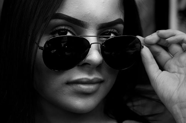 A woman poses with aviator sunglasses in a striking, high-contrast black and white portrait.