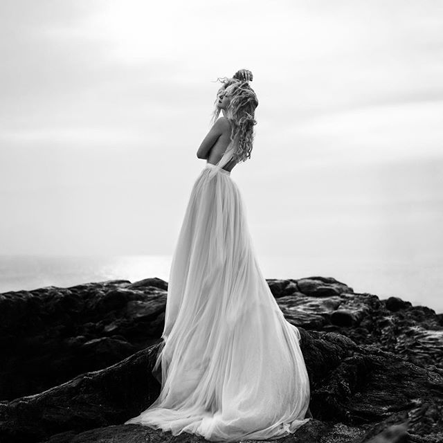 A woman in a flowing dress stands on a rocky coast, looking out at the ocean in a serene, black and white editorial shot.