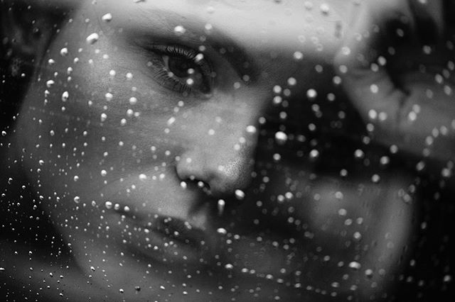 A grayscale portrait of a woman's face with raindrops on a window, conveying a sense of intimacy and emotion.
