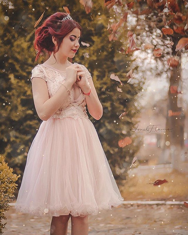 A young woman in a pink dress stands serenely in a park with falling leaves.