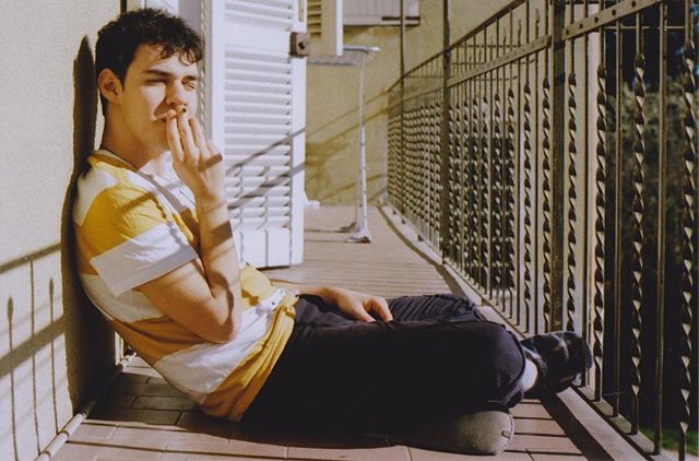 A young man sits on a sunlit balcony, pausing to smoke a cigarette in a moment of contemplation.