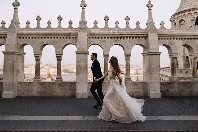 A bride and groom hold hands, running in front of an ornate stone building with a cityscape view.
