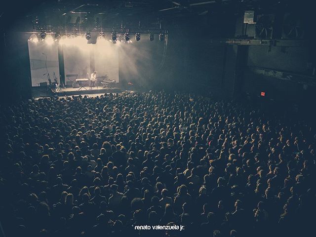 A large crowd enjoys a live music concert with a performer on stage highlighted by spotlights in a dimly lit venue.