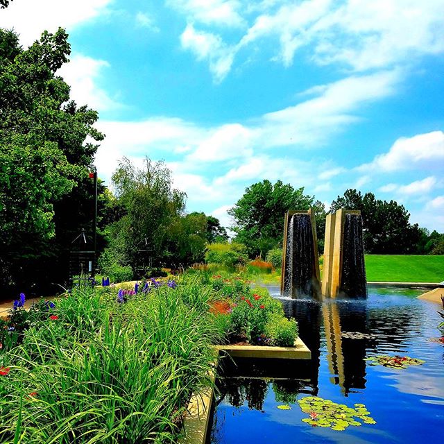 A tranquil garden scene with a fountain, pond, and vibrant greenery under a blue sky, perfect for relaxation and escape.