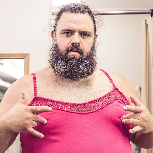 A bearded man playfully poses in a pink dress, challenging gender norms with humor.