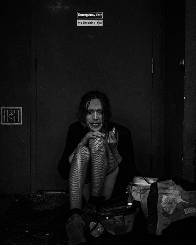 A woman in fishnet stockings sits in a doorway under an emergency exit and no smoking sign in a moody, black and white urban scene.