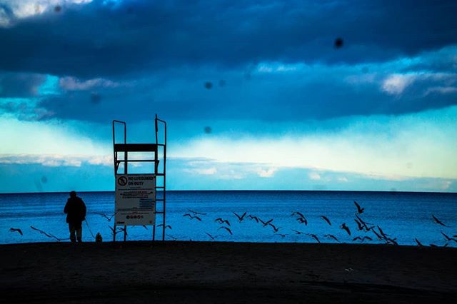 A solitary figure stands on a beach next to a lifeguard chair as birds fly over the calm ocean.